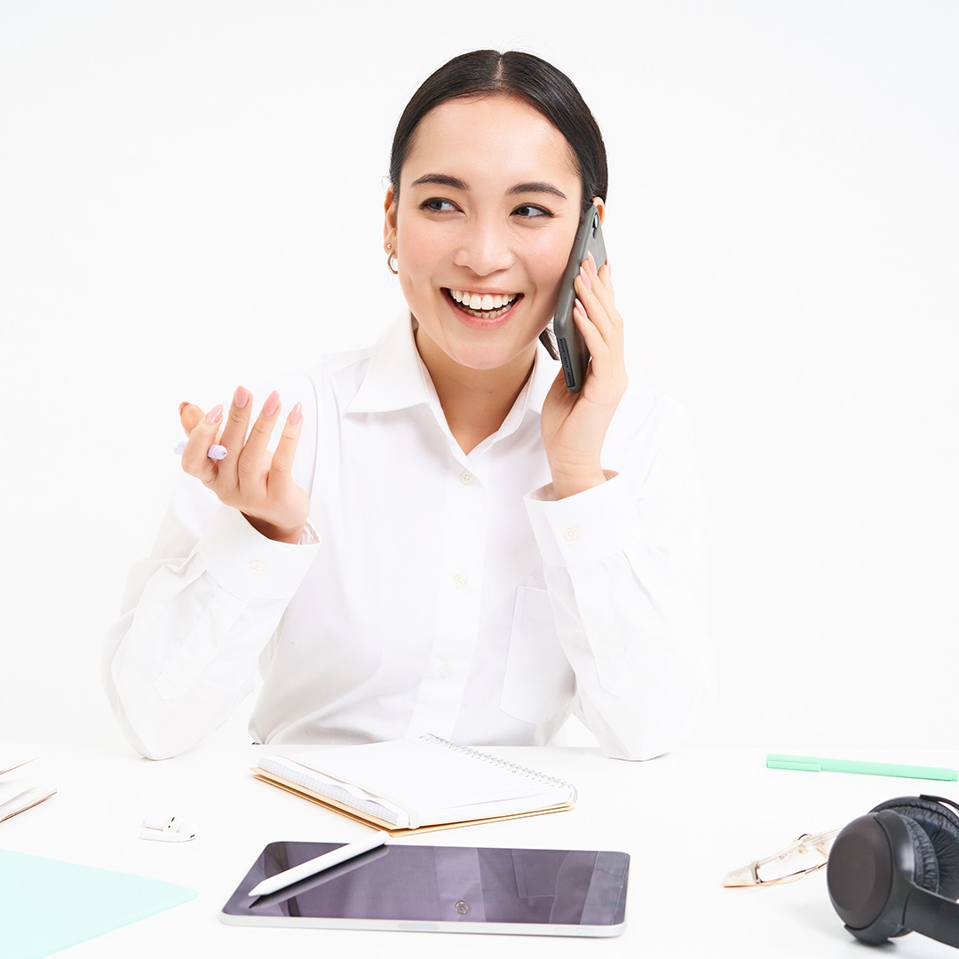A smiling female healthcare representative in a white shirt, talking on the phone at a desk with a notebook, tablet, and office supplies.